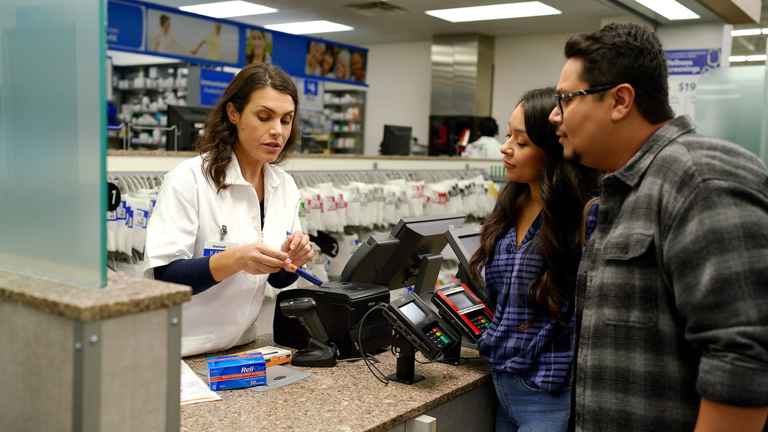 A female Walmart Pharmacist stands behind the counter holding a ReliOn insulin pen while pointing to the label. She is seen discussing with male and female customers.