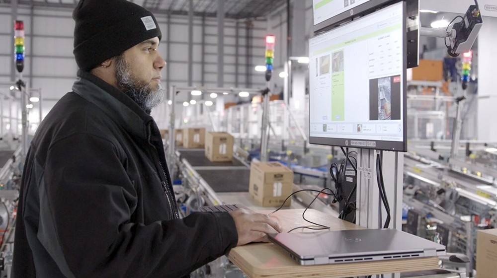 An associate wearing a black jacket and white beanie faces a computer monitor. He is using a laptop placed on a table to his right. There is a conveyor belt with boxes on it behind him.