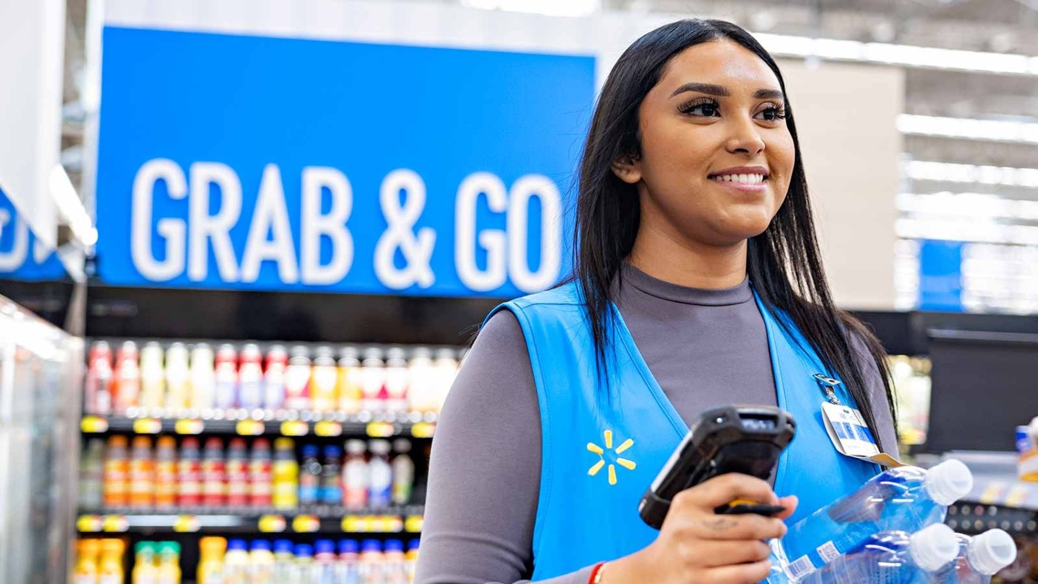 A Walmart associate stands in the Grab & Go section with a handheld scanner and water bottles in her hand.