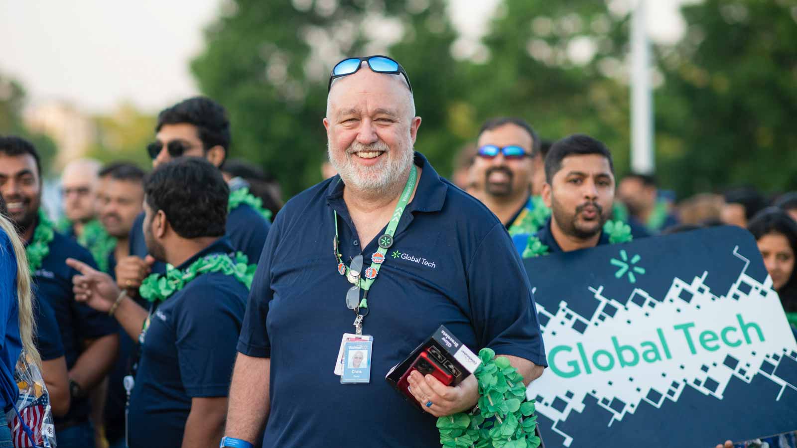 Associate on the Global Tech team is seen smiling at the camera. He fellow Global Tech associates are seen in the background with their sign that reads "Global Tech".