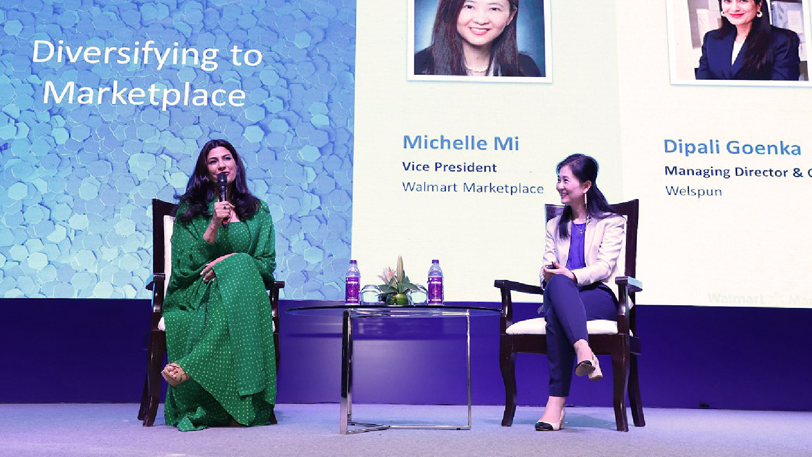 Indian woman in green sari, speaking into a microphone. On right sits an Asian American woman, listening attentively. Background reads: Diversifying the Marketplace.